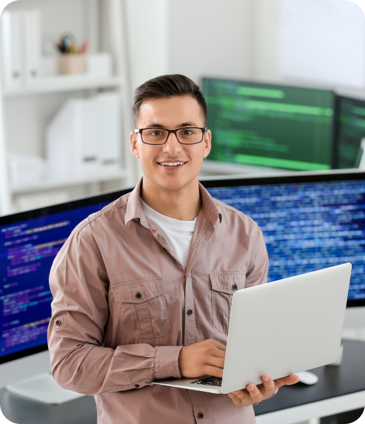 A man with glasses holding a laptop in front of monitors with software development code in the background