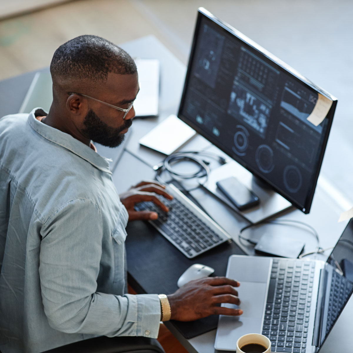 Man working at desk with multiple screens, using laptop and keyboard, focusing on data analysis.