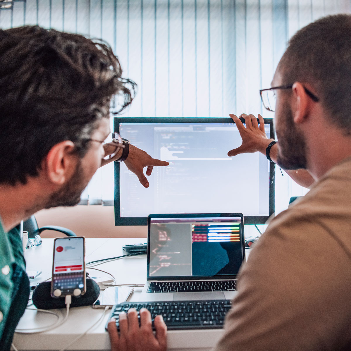Two individuals collaborate on coding project with multiple screens and a smartphone, working closely at a computer desk.