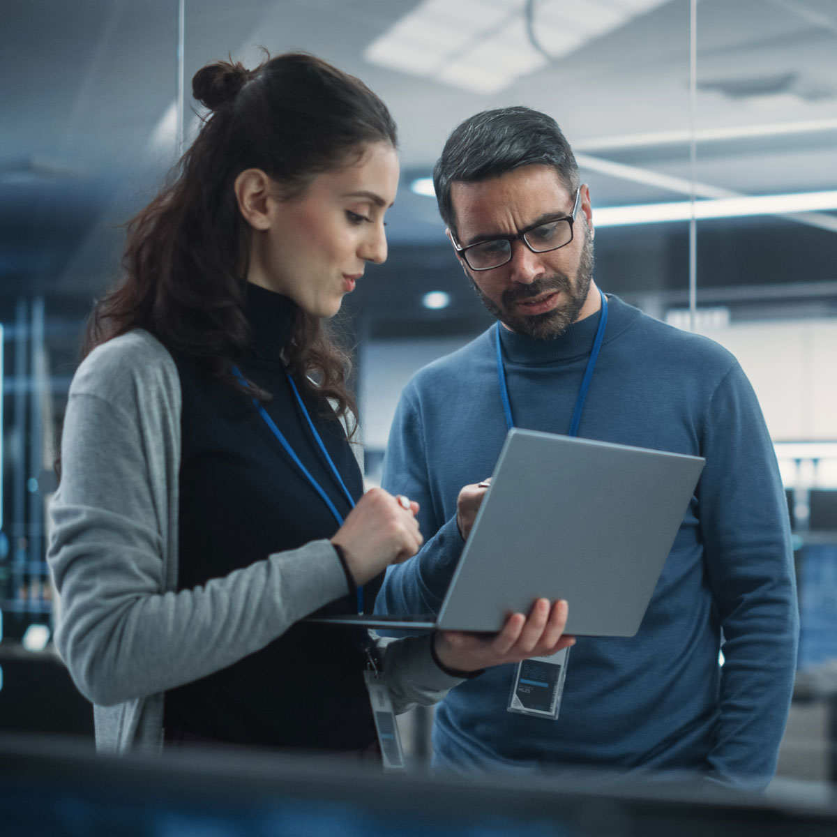 Two professionals with lanyards discuss work while looking at a laptop in a modern office setting.