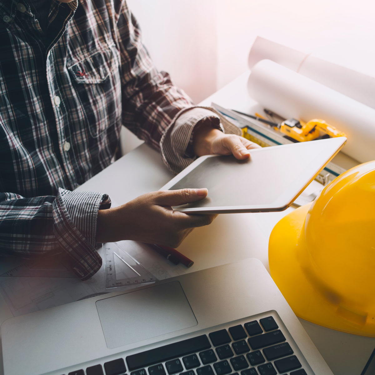 AEC professional holding a tablet with a laptop and a yellow hard hat on the table