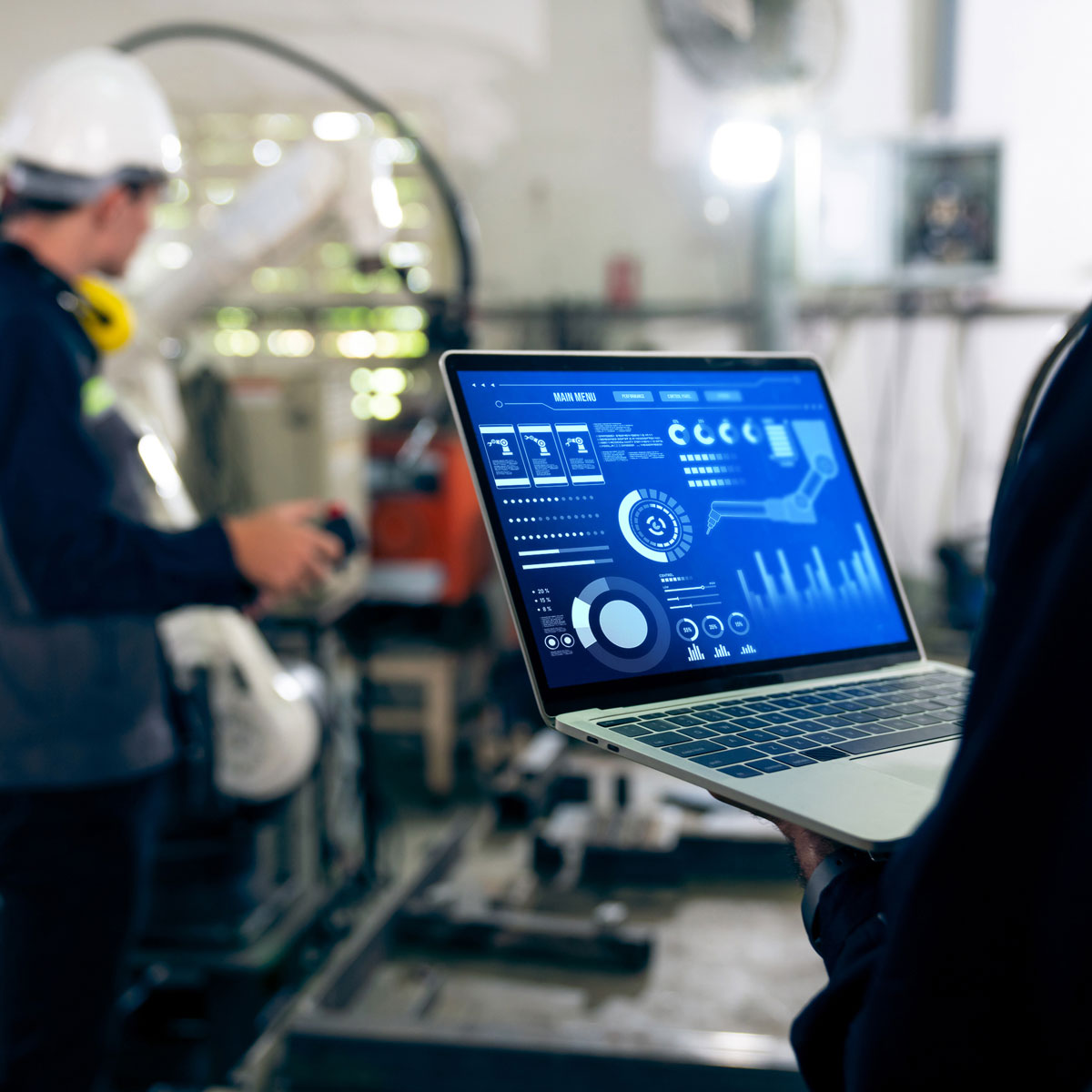 A person holding a laptop showing metrics with engineering equipment in the background
